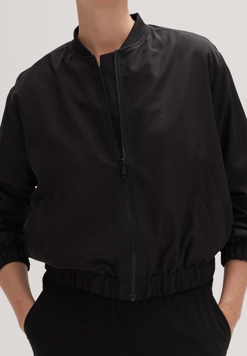Czarna kurtka bomberka wykonana z gładkiej tkaniny. Posiada zamek błyskawiczny z przodu, elastyczne mankiety i pas biodrowy. Brak widocznych wzorów czy elementów metalowych.