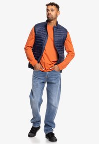 Chaleco acolchado azul marino sobre una camiseta de manga larga naranja, combinado con unos jeans azules y zapatillas negras. El chaleco tiene un cuello alto y costuras verticales.