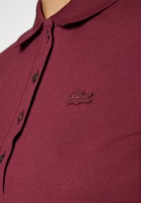 Gros plan sur un polo bordeaux à boutons avec un logo crocodile brodé sur la poitrine gauche.