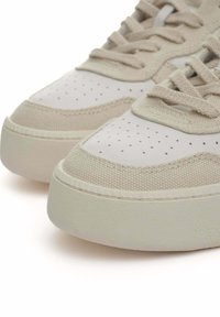 Sneakers beige con una toma superiore in tessuto texture, punta in pelle liscia e dettagli perforati. Suola in gomma spessa con una leggera lucentezza.