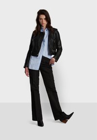 Black leather cropped jacket, light blue button-up shirt, and black flared trousers. Model wears black heeled ankle boots. Simple background.