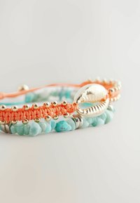 Multistrenget armbånd med en sølvskallaccent, koraltråd og turkise stenperler, som fremviser en blanding af teksturer og farver.