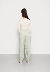Femme avec de longs cheveux tressés portant un haut ajusté à manches longues et un pantalon en satin vert pâle ample, se tenant face à un mur blanc.