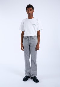Camiseta blanca de algodón con logo verde, combinada con vaqueros acampanados de color gris claro. El modelo lleva zapatos de plataforma negros. Conjunto sencillo y casual.