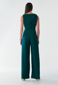 Teal satin jumpsuit med vidde ben, ærmeløs krop og ryglynlås. Har diskret plissering ved skuldrene og rene linjer.