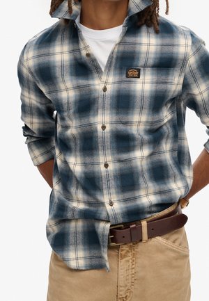 Mann trägt blau-weiß kariertes Hemd mit Knöpfen über weißem T-Shirt, beige Hose mit braunem Gürtel, Hände hinter dem Rücken.