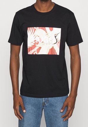 Mężczyzna w czarnej koszulce z grafiką białego żurawia na czerwonym, abstrakcyjnym tle, ubrany w niebieskie dżinsy, stojący na jednolitym tle.