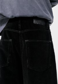Pantaloni din corduroy negri cu rigle verticale pronunțate, două buzunare la spate cu cusături albe și o etichetă din piele neagră pe talie.