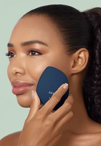 Dispositif de nettoyage facial bleu aux bords arrondis, tenu contre la peau. Les boutons pour les réglages sont visibles. Texture lisse et design ergonomique.