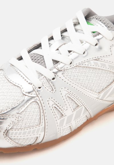 Gros plan d'une basket argentée et grise avec des lacets blancs et une semelle marron clair sur fond blanc.