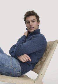 Dunkelblauer Strickpullover mit hohem Kragen, kombiniert mit hellblauen Jeans, sitzt auf einem Holzstuhl mit geflochtenem Muster.