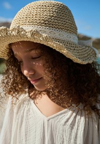Chapeau en paille à larges bords avec un tissage texturé et un bord froncé, doté d'un bandeau en dentelle blanche. Une lumière douce crée des motifs d'ombre.