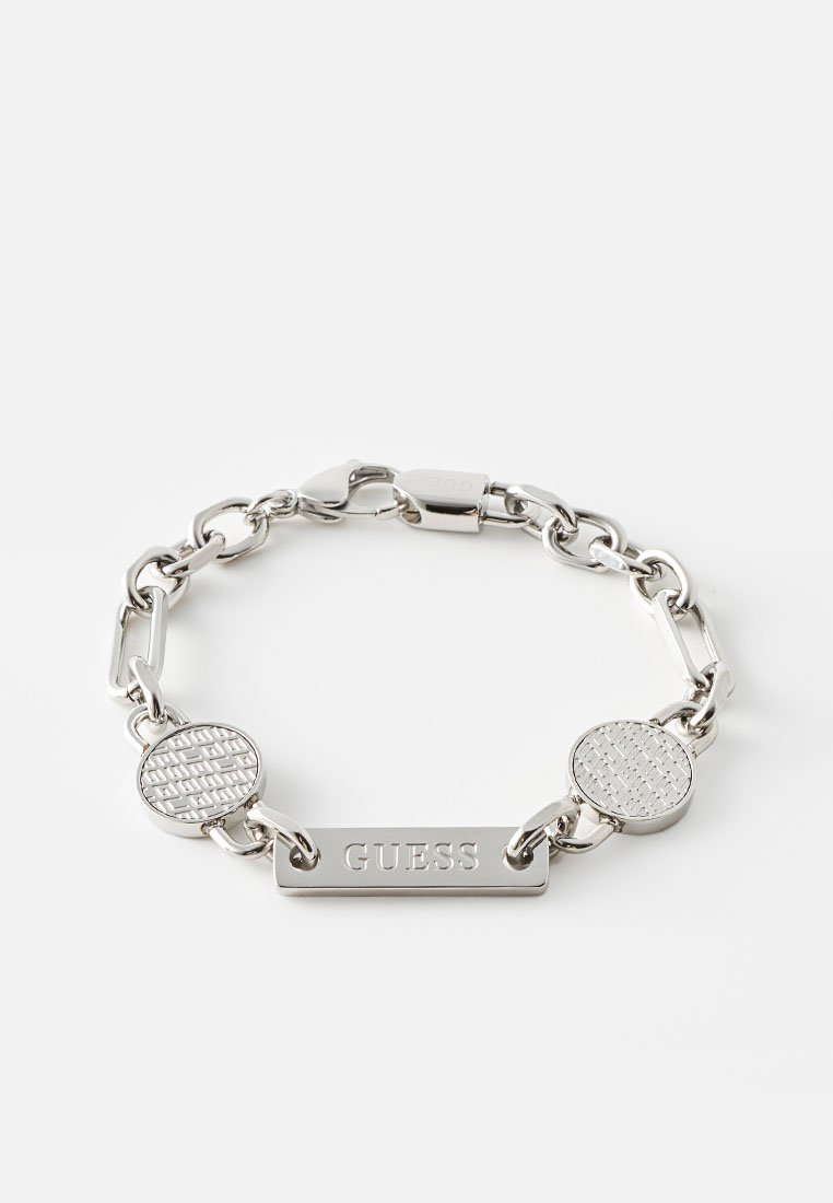 Bracelet en chaîne argentée avec des maillons circulaires texturés et une étiquette rectangulaire centrale gravée "GUESS". Fini métallique poli.