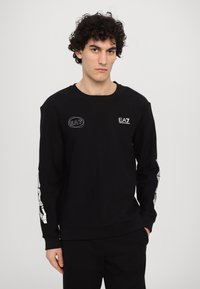 Črna crew-neck majica s teksturirano tkanino, ki ima izvezen EA7 logotip na prsih in grafičen detalj ob rokavih.