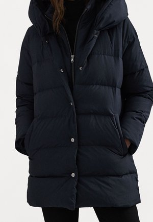 Femme portant une doudoune noire matelassée avec une fermeture éclair et des boutons-pression, des poches latérales et un col montant avec une capuche.