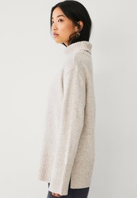 Maglione a collo alto di un beige chiaro, realizzato in un morbido tessuto testurizzato. Presenta polsini e orlo a costine, con una vestibilità rilassata e cuciture delle spalle scese.