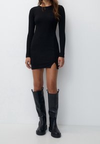 Mini-robe noire à manches longues avec une fente latérale, confectionnée dans un tissu lisse. Associée à des bottes en cuir noires hauteur genou avec semelles épaisses.