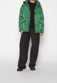 Veste à capuche verte en duvet, dotée d'un design matelassé, d'une fermeture à pression et de poches avant. Portée par-dessus un pull noir et un pantalon ample.