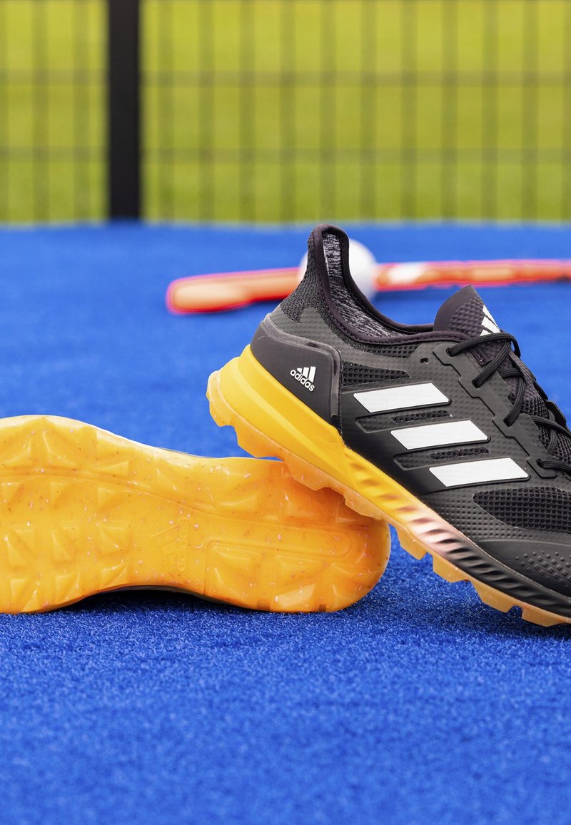 Chaussure de sport noire avec trois rayures blanches, dotée d'un dessus texturé et d'une semelle en caoutchouc orange-jaune avec un motif de bande de roulement.