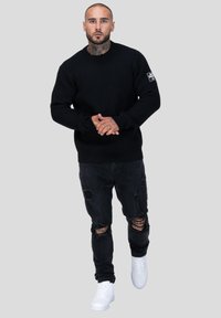 Zwarte ribgebreide trui met lange mouwen, gecombineerd met versleten zwarte jeans en witte sneakers. Geen logo's of andere patronen zichtbaar.