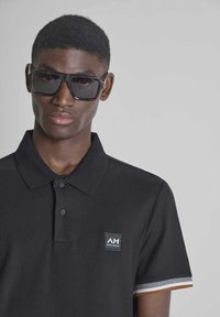 Joven con gafas de sol oscuras y una camisa polo negra con un pequeño logo en el pecho izquierdo, sobre un fondo liso.