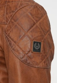 Καφέ δερμάτινο μπουφάν με ραμμένη λεπτομέρεια στους ώμους, που διαθέτει μαύρο λογότυπο Belstaff από καουτσούκ και υφή στην επιφάνεια.