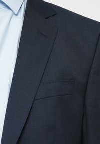 Marinblå kostymjacka med en texturerad tyg, med en notch-lapel och en enkel bröstficka. Ljusblå skjorta synlig under.