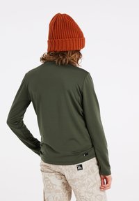 Grünes Langarmoberteil mit glatter Textur, kombiniert mit hellbeigen Hosen mit gemustertem Design und einem schwarzen Logoetikett.