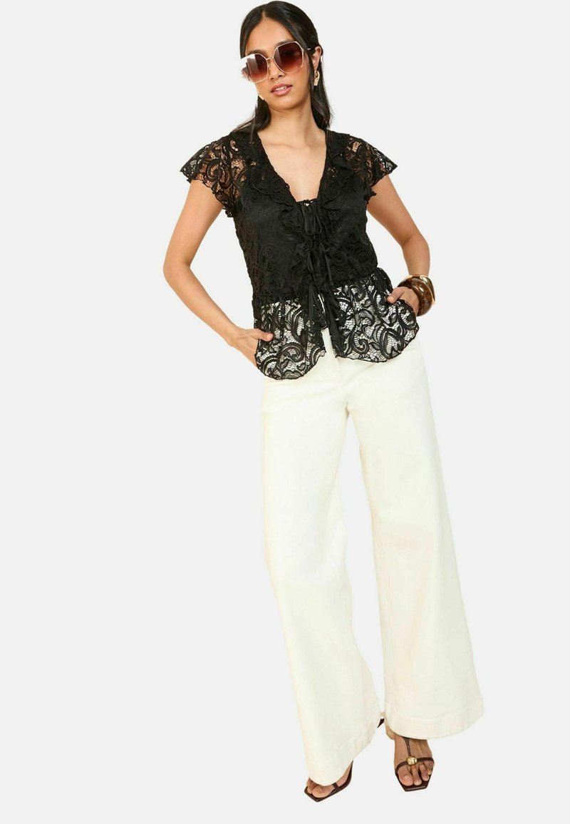 Top in pizzo nero con motivi floreali, maniche corte a volant e chiusura con cravatta, abbinato a pantaloni larghi ivory e sandali neri.