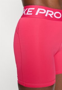 Ryškiai rožinės Nike Pro kompresinės šortai su plačiu kontrastiniu juosmeniu, ant kurio yra užrašas "NIKE PRO". Sklandi tekstūra ir prigludusi forma.