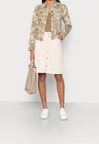 Chaqueta de mezclilla ligera color canela con patrón de camuflaje sobre una blusa marrón, combinada con una falda beige con botones y zapatillas blancas. Lleva una bolsa tote beige en la mano.