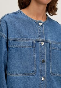 Hellblaue Jeansjacke mit zwei großen Fronttaschen. Verfügt über silberne Druckknöpfe, einen runden Ausschnitt und sichtbare Nähte.