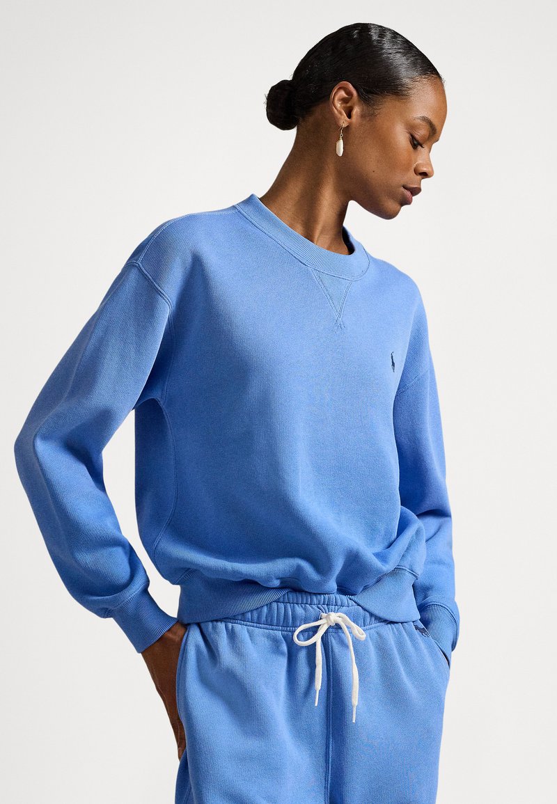 Sweatshirt azul claro em tecido macio, com gola redonda canelada, mangas compridas e um pequeno logótipo no peito. Combinado com calças à altura.