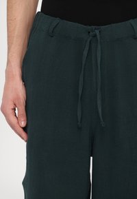 Dark green linen trousers with a drawstring waist, featuring a textured finish and side pockets. Hand rests on the waistband.