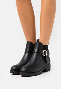 Bottines noires en cuir lisse avec une sangle texturée et une boucle en argent. Semelle épaisse avec un motif pour une adhérence optimale.