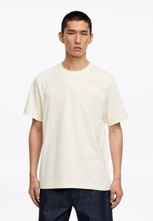 Jeune homme debout, portant un t-shirt à manches courtes uni blanc cassé et un pantalon foncé, sur fond blanc.