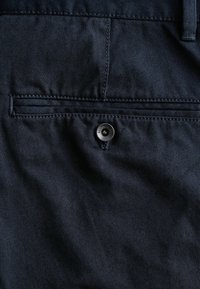 Tissu en denim bleu foncé au toucher lisse, avec une poche arrière ornée d'un bouton en métal noir visible.