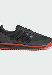 Baskets noires avec une tige en suède texturé et synthétique, trois bandes noires, et une semelle en caoutchouc rouge à motif en zigzag.