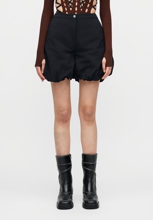 Άτομο που φοράει μαύρα φουσκωτά σορτς, μαύρες μπότες μέχρι τη μέση της γάμπας με λευκή ραφή και καφέ μακρυμάνικη μπλούζα με ανοιχτά σημεία για τους αντίχειρες.