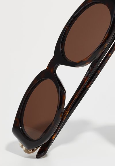 Occhiali da sole tartarugati con lenti rotonde marroni e una montatura spessa. Le aste presentano un design elegante con un sottile dettaglio di branding.
