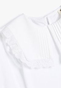Weiße Bluse mit plissierter Vorderseite und einem gewellten, bestickten Spitzenkragen. Glattes Material mit einem sauberen Ausschnitt und Rückenverschluss.