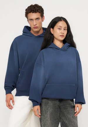 To unge voksne iført blå hættetrøjer, den ene med hvide bukser og den anden med grå jeans, der står roligt mod en ensfarvet baggrund.