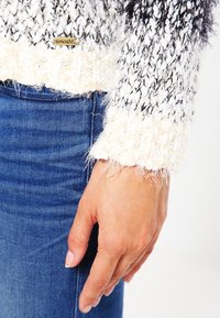 Cremefarbener und schwarzer Strickpullover mit flauschiger Textur, ausgestattet mit einem gerippten Abschluss und eng anliegenden Bündchen. Goldenes Logo-Detail am Saum. Kombiniert mit blauen Jeans.