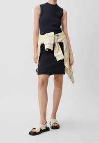 Robe côtelée bleu marine avec un col montant, accompagnée d'une étole rayée jaune clair. La personne porte des sandales à lanières croisées blanches et noires.