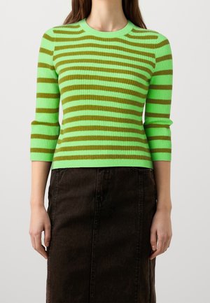 Eine Frau trägt einen grün-braunen horizontal gestreiften Ripp-Pullover mit Dreiviertelärmeln und einen dunkelbraunen Rock.