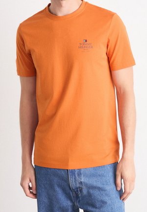 Orange cotton t-shirt with short sleeves, round neckline, and a small navy logo on the left chest; paired with blue jeans.