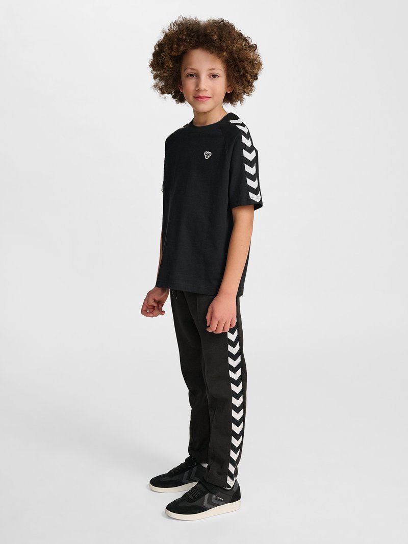 Dziecko z kręconymi włosami w czarnej koszulce i spodniach z białymi chevronowymi paskami na rękawach i nogawkach, stojące bokiem.