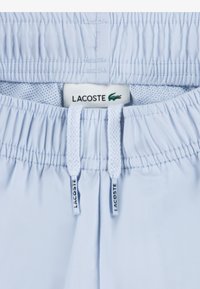 Ανοιχτό μπλε μαγιό σορτς με κορδόνι στη μέση, με υφασμένη ελαστική ζώνη και ετικέτα με το λογότυπο Lacoste ορατή στη μέση.