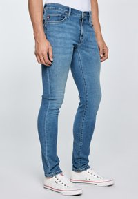 Mees, kes kannab helesiniseid kitsaid teksaseid ja valgeid madala säärega tenniseid, seisab ühevärvilise tausta ees.