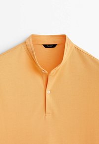 Oranges Poloshirt mit Stehkragen, zwei Knöpfen und geripptem Material. Das Etikett im Kragen zeigt ein schwarz-weißes Branding.